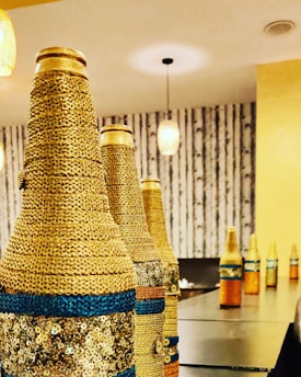 Decorative bottles wrapped in gold and blue materials are arranged in a row on a table. The setting includes warm lighting and a patterned wall background, creating an elegant and cozy atmosphere.