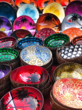 A vibrant assortment of hand-painted bowls, each featuring intricate designs and bright colors. The bowls display a variety of patterns, including floral motifs and scenic imagery with palm trees. The backgrounds are painted in rich shades of red, blue, green, purple, and orange, with some bowls having a glossy finish.