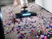 A vacuum cleaner is being used to clean colorful confetti off a grey carpet. The vacuum is silver with a lit nozzle showing the confetti being sucked in. A pair of bare feet is visible behind the device, indicating a person is operating it.