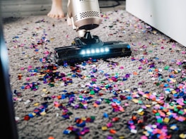 A vacuum cleaner is being used to clean colorful confetti off a grey carpet. The vacuum is silver with a lit nozzle showing the confetti being sucked in. A pair of bare feet is visible behind the device, indicating a person is operating it.