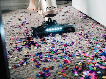 A vacuum cleaner is being used to clean colorful confetti off a grey carpet. The vacuum is silver with a lit nozzle showing the confetti being sucked in. A pair of bare feet is visible behind the device, indicating a person is operating it.