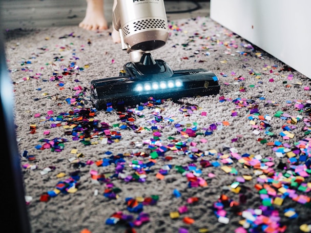 A vacuum cleaner is being used to clean colorful confetti off a grey carpet. The vacuum is silver with a lit nozzle showing the confetti being sucked in. A pair of bare feet is visible behind the device, indicating a person is operating it.