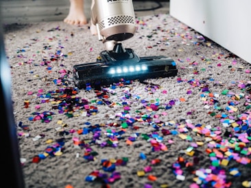 A vacuum cleaner is being used to clean colorful confetti off a grey carpet. The vacuum is silver with a lit nozzle showing the confetti being sucked in. A pair of bare feet is visible behind the device, indicating a person is operating it.