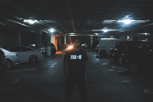 A dimly lit parking garage with several parked cars. A person stands in the foreground wearing a shirt with a white text that reads 'JESUS > ME'. Overhead lights provide sparse illumination, creating a moody and shadow-filled atmosphere.