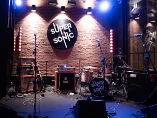 A music venue stage setup with various musical equipment, including drums, guitars, amplifiers, and microphones. The brick wall backdrop features a large circular 'SUPER SONIC' sign illuminated by overhead spotlights, creating a warm and inviting atmosphere. The scene is ready for a live performance.