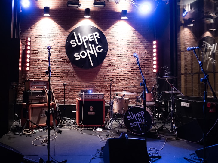 A music venue stage setup with various musical equipment, including drums, guitars, amplifiers, and microphones. The brick wall backdrop features a large circular 'SUPER SONIC' sign illuminated by overhead spotlights, creating a warm and inviting atmosphere. The scene is ready for a live performance.
