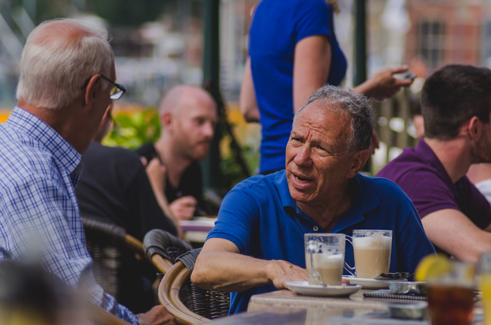 青いポロシャツを着た男がコーヒーを飲みながら男に話しかける