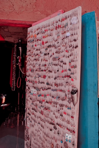A display board adorned with a wide variety of colorful earrings, showcasing numerous styles and designs. The earrings are neatly arranged in rows, with each pair featuring different colors, shapes, and materials. The backdrop includes a textured wall with some additional hanging jewelry items.