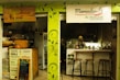 A small, cozy café features a wooden counter and shelves filled with organic products. The café's signage emphasizes natural and intelligent cooking. A person wearing a headscarf stands behind the counter, with stools lined up for customers.