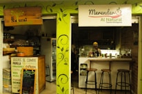 A small, cozy café features a wooden counter and shelves filled with organic products. The café's signage emphasizes natural and intelligent cooking. A person wearing a headscarf stands behind the counter, with stools lined up for customers.