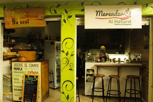 A small, cozy café features a wooden counter and shelves filled with organic products. The café's signage emphasizes natural and intelligent cooking. A person wearing a headscarf stands behind the counter, with stools lined up for customers.