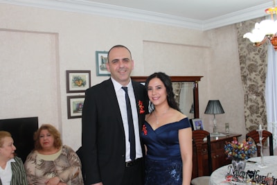 A man in a black suit and a woman in a navy blue dress stand smiling in a living room. Behind them, two older women are seated. The room is decorated with framed pictures on the wall, a table with a lamp, flowers, and a chandelier. The furniture and curtains have a classic style.