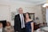 A man in a black suit and a woman in a navy blue dress stand smiling in a living room. Behind them, two older women are seated. The room is decorated with framed pictures on the wall, a table with a lamp, flowers, and a chandelier. The furniture and curtains have a classic style.
