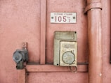 An old emergency phone mounted on a textured pink wall. It features a worn-out rectangular housing with an instruction label in French. Nearby, a metal pipe runs vertically alongside the wall.
