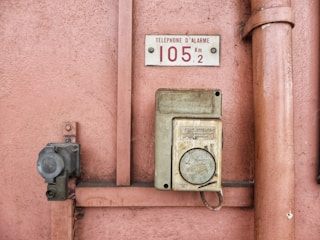 An old emergency phone mounted on a textured pink wall. It features a worn-out rectangular housing with an instruction label in French. Nearby, a metal pipe runs vertically alongside the wall.