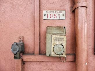 An old emergency phone mounted on a textured pink wall. It features a worn-out rectangular housing with an instruction label in French. Nearby, a metal pipe runs vertically alongside the wall.