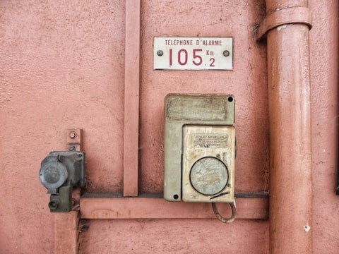 An old emergency phone mounted on a textured pink wall. It features a worn-out rectangular housing with an instruction label in French. Nearby, a metal pipe runs vertically alongside the wall.