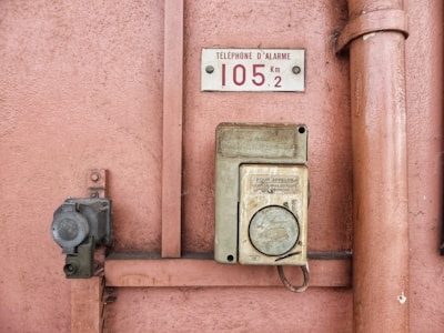 An old emergency phone mounted on a textured pink wall. It features a worn-out rectangular housing with an instruction label in French. Nearby, a metal pipe runs vertically alongside the wall.