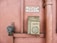 An old emergency phone mounted on a textured pink wall. It features a worn-out rectangular housing with an instruction label in French. Nearby, a metal pipe runs vertically alongside the wall.