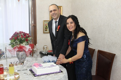 A couple is standing behind a table, smiling and cutting a white cake with purple decorations. The woman is dressed in a blue dress, while the man is wearing a black suit with a white shirt. The table is adorned with various items, including candles, beverages, and a bouquet of red roses wrapped in white and red paper. The room has light-colored walls and framed pictures hanging in the background.
