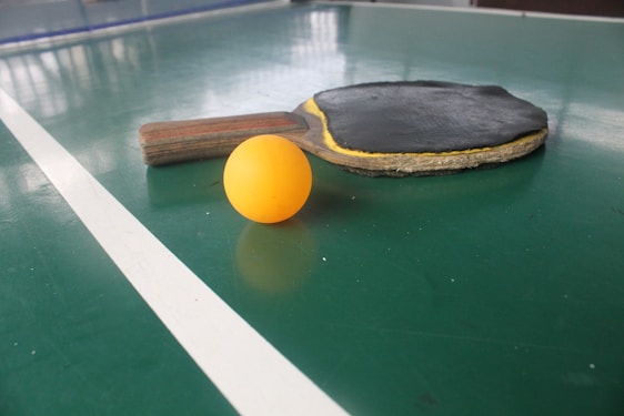 A table tennis paddle with a black rubber surface is laid next to an orange ping pong ball on a green table with white lines marking the surface.