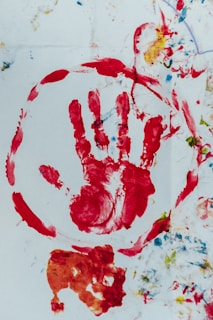 A red handprint is encircled in red paint on a white background, surrounded by various splashes of color including blue and yellow. There are additional abstract forms and smudges below the handprint in orange and red hues.