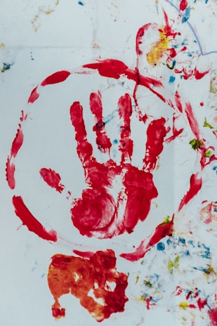 A red handprint is encircled in red paint on a white background, surrounded by various splashes of color including blue and yellow. There are additional abstract forms and smudges below the handprint in orange and red hues.