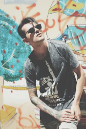 A model wearing a graphic tee with vibrant favela art, standing near a graffiti-covered wall.
