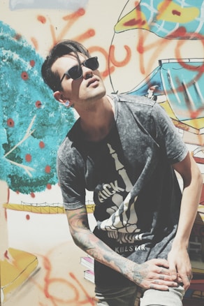 A person wearing sunglasses and a graphic t-shirt poses in front of a colorful graffiti wall. The mural includes abstract shapes and bold, bright colors. The individual&rsquo;s arms are tattooed, and they lean slightly with an upward gaze.