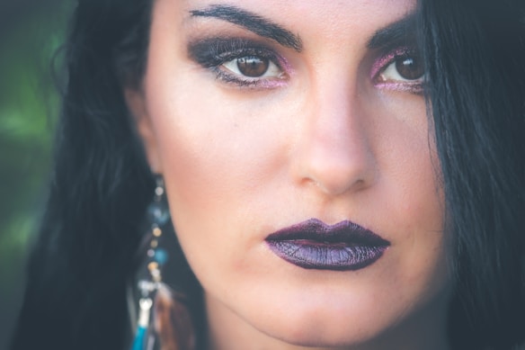 A close-up portrait of a person with striking dark makeup, including bold eyeliner and dark lipstick. The focus is on the eyes and lips, with long dark hair framing the face. An earring with feathers and beads is partially visible.
