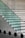 A modern stairway with glass steps and a metal railing. The steps transition from a green tint to a more transparent appearance as they ascend. The staircase is set against a concrete wall, creating a minimalist and sleek design. There are black circular shadows or shapes at the bottom, suggesting an artistic or architectural element.