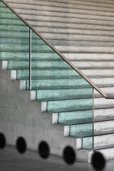 A modern stairway with glass steps and a metal railing. The steps transition from a green tint to a more transparent appearance as they ascend. The staircase is set against a concrete wall, creating a minimalist and sleek design. There are black circular shadows or shapes at the bottom, suggesting an artistic or architectural element.