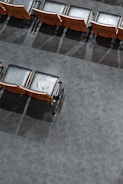Rows of empty, black cushioned seats with wooden backs are neatly aligned on a gray carpeted floor. The seats' shadows are cast on the floor, and there is an orderly, symmetrical pattern to their arrangement.