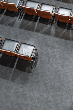 Rows of empty, black cushioned seats with wooden backs are neatly aligned on a gray carpeted floor. The seats' shadows are cast on the floor, and there is an orderly, symmetrical pattern to their arrangement.