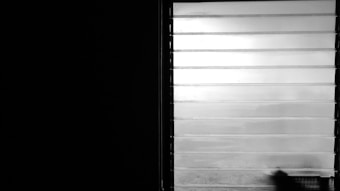 A window with horizontal blinds slightly opened allows diffused light to enter a dimly lit room. The left side of the image is dominated by darkness, contrasting sharply with the bright window. A blurred, indistinct shadow or object appears at the bottom right corner of the window.