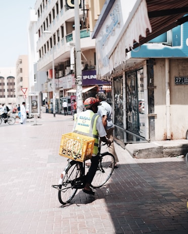 A person wearing a bright safety vest and a helmet rides a bicycle down a city street. The bicycle has a large yellow crate attached to the back. The street is lined with a variety of shops and buildings, and there are other pedestrians in the background. The atmosphere is busy with people and vehicles, typical of an urban environment.