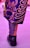 A close-up of a person's lower leg and foot wearing a brightly patterned garment with swirling designs in purple and gold colors, paired with a black Nike Air shoe. The ground has a pinkish hue, reflecting a vivid color scheme.
