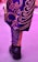 A close-up of a person's lower leg and foot wearing a brightly patterned garment with swirling designs in purple and gold colors, paired with a black Nike Air shoe. The ground has a pinkish hue, reflecting a vivid color scheme.