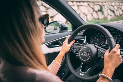 A person is driving a car, holding the steering wheel with both hands. The interior of the vehicle, specifically the dashboard and steering controls, is visible. The car seems to be an Audi, as indicated by the logo on the steering wheel. The driver is wearing sunglasses and a sweater, and the road is visible through the windows.