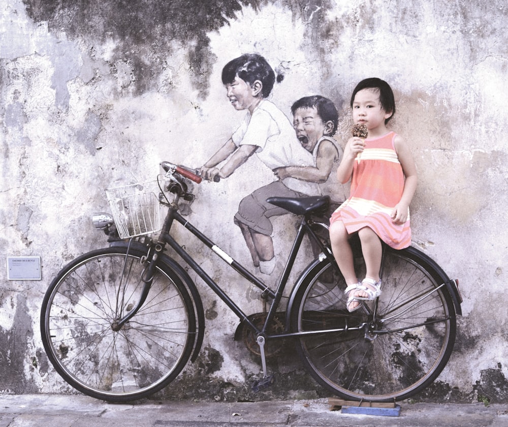 girl wearing orange and white sleeveless dress eating ice cream while sitting on black and white bike leaning on wall