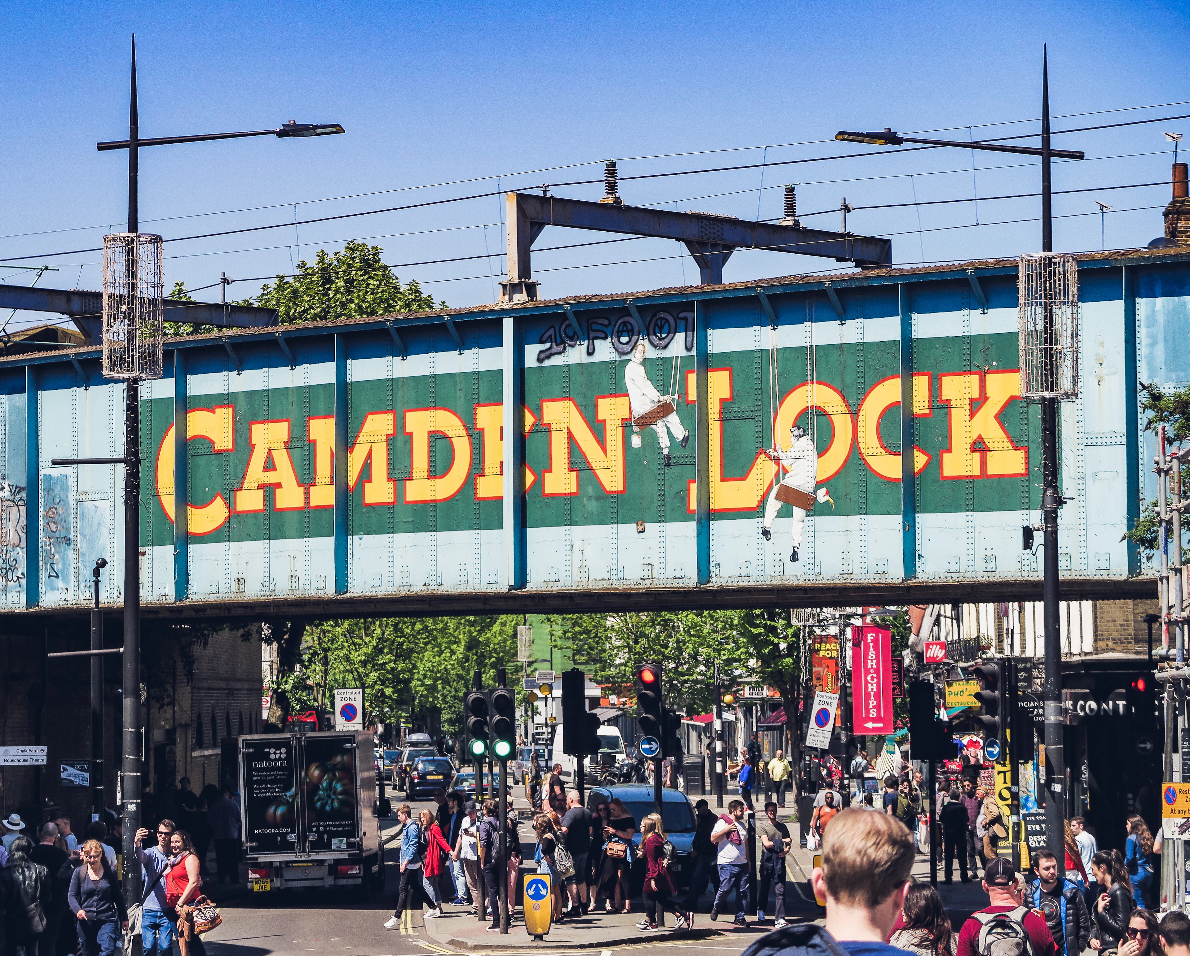 Crowds passing beneath the green railway bridge lettered CAMDEN LOCK at Camden Market, London, on a sunny day