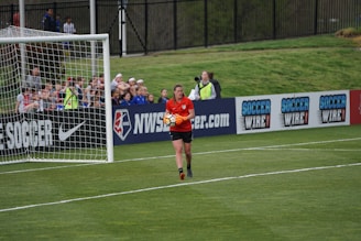 A soccer player wearing a red jersey holds a soccer ball near the goalpost on a well-maintained grass field. In the background, spectators are sitting and watching the game, while advertisements for soccer-related products are displayed along the sideline fences.