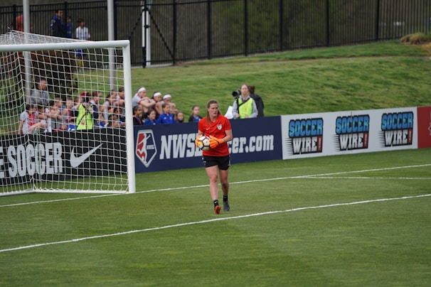 A soccer player wearing a red jersey holds a soccer ball near the goalpost on a well-maintained grass field. In the background, spectators are sitting and watching the game, while advertisements for soccer-related products are displayed along the sideline fences.