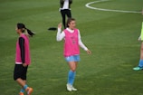A group of soccer players on a grassy field wearing pink training vests. One player is gesturing with her hand while another stands nearby with her hands on her hips. They are wearing shorts and long socks, and the field is marked with white lines.