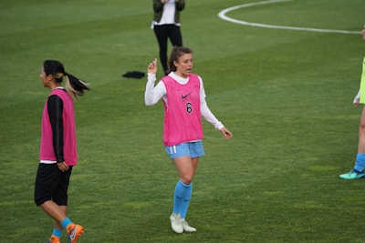 A group of soccer players on a grassy field wearing pink training vests. One player is gesturing with her hand while another stands nearby with her hands on her hips. They are wearing shorts and long socks, and the field is marked with white lines.