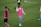 A group of soccer players on a grassy field wearing pink training vests. One player is gesturing with her hand while another stands nearby with her hands on her hips. They are wearing shorts and long socks, and the field is marked with white lines.