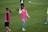 A group of soccer players on a grassy field wearing pink training vests. One player is gesturing with her hand while another stands nearby with her hands on her hips. They are wearing shorts and long socks, and the field is marked with white lines.