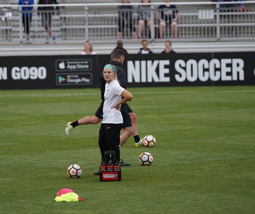 A person in sports attire stands on a soccer field, surrounded by several soccer balls and a carrier with bottles. The field is bordered by advertisements for Nike Soccer and app stores. Several people sit in bleachers in the background.