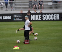 A person in sports attire stands on a soccer field, surrounded by several soccer balls and a carrier with bottles. The field is bordered by advertisements for Nike Soccer and app stores. Several people sit in bleachers in the background.