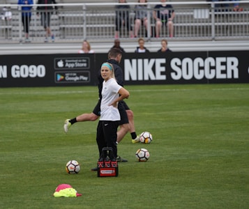 A person in sports attire stands on a soccer field, surrounded by several soccer balls and a carrier with bottles. The field is bordered by advertisements for Nike Soccer and app stores. Several people sit in bleachers in the background.